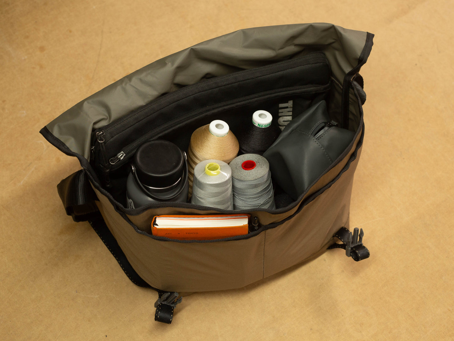Top-down view of packed wipwrk Mondo messenger bag in brown with water-resistant lining, filled with sewing thread spools, reusable water bottle, zippered organizer pouch, and notebook