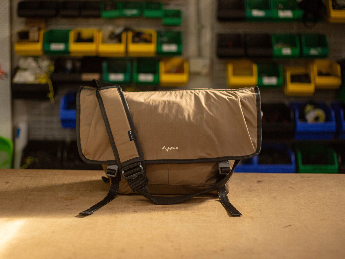 Front view of wipwrk Mondo messenger bag in brown showing durable exterior, adjustable padded shoulder strap, and quick-release buckles against a workshop backdrop