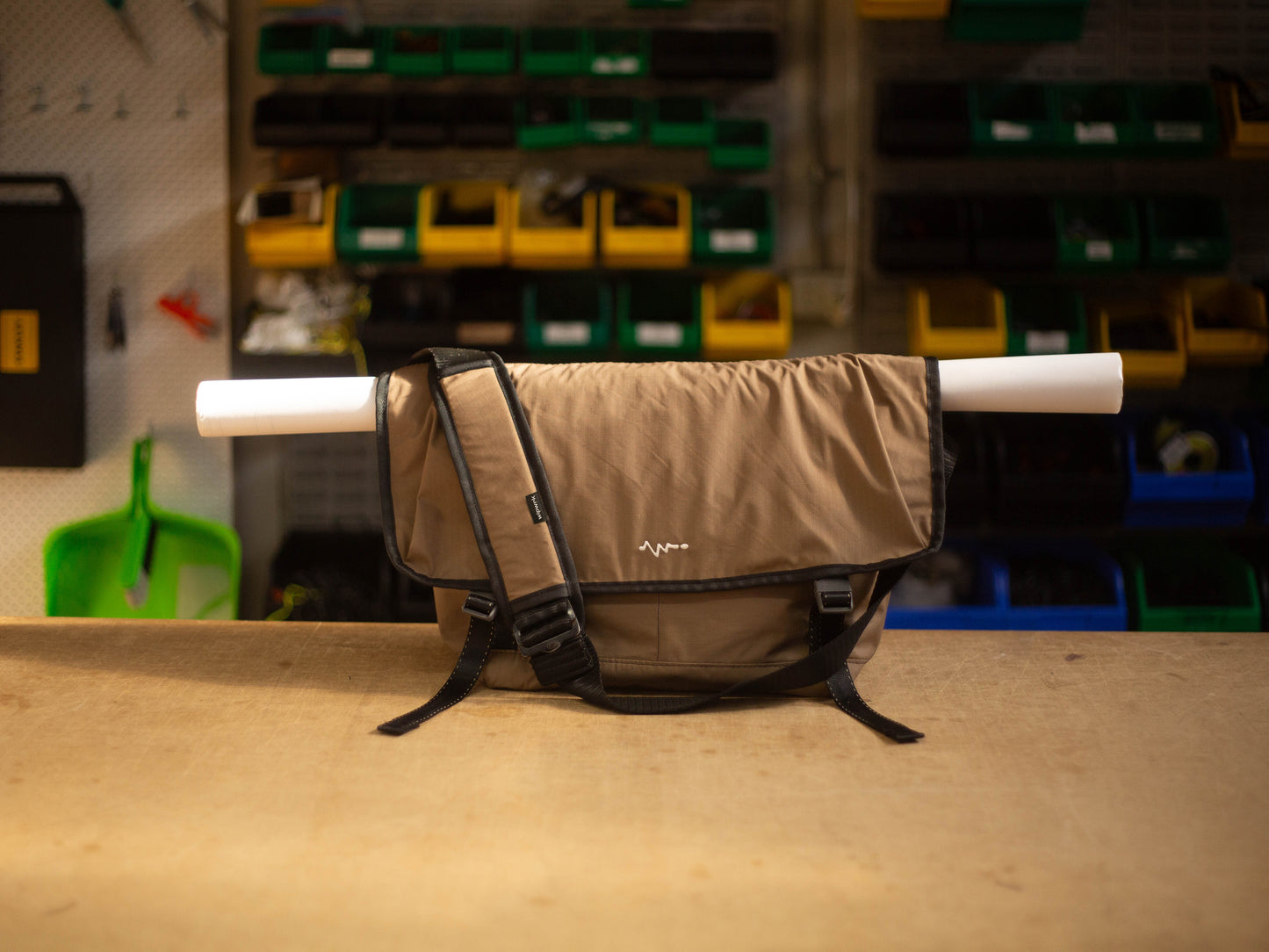 Hero shot of wipwrk Mondo Hauler messenger bag in brown with top flap rolled up and secured by integrated straps carrying a rolled document tube, showcasing adjustable padded shoulder strap and quick-release buckles