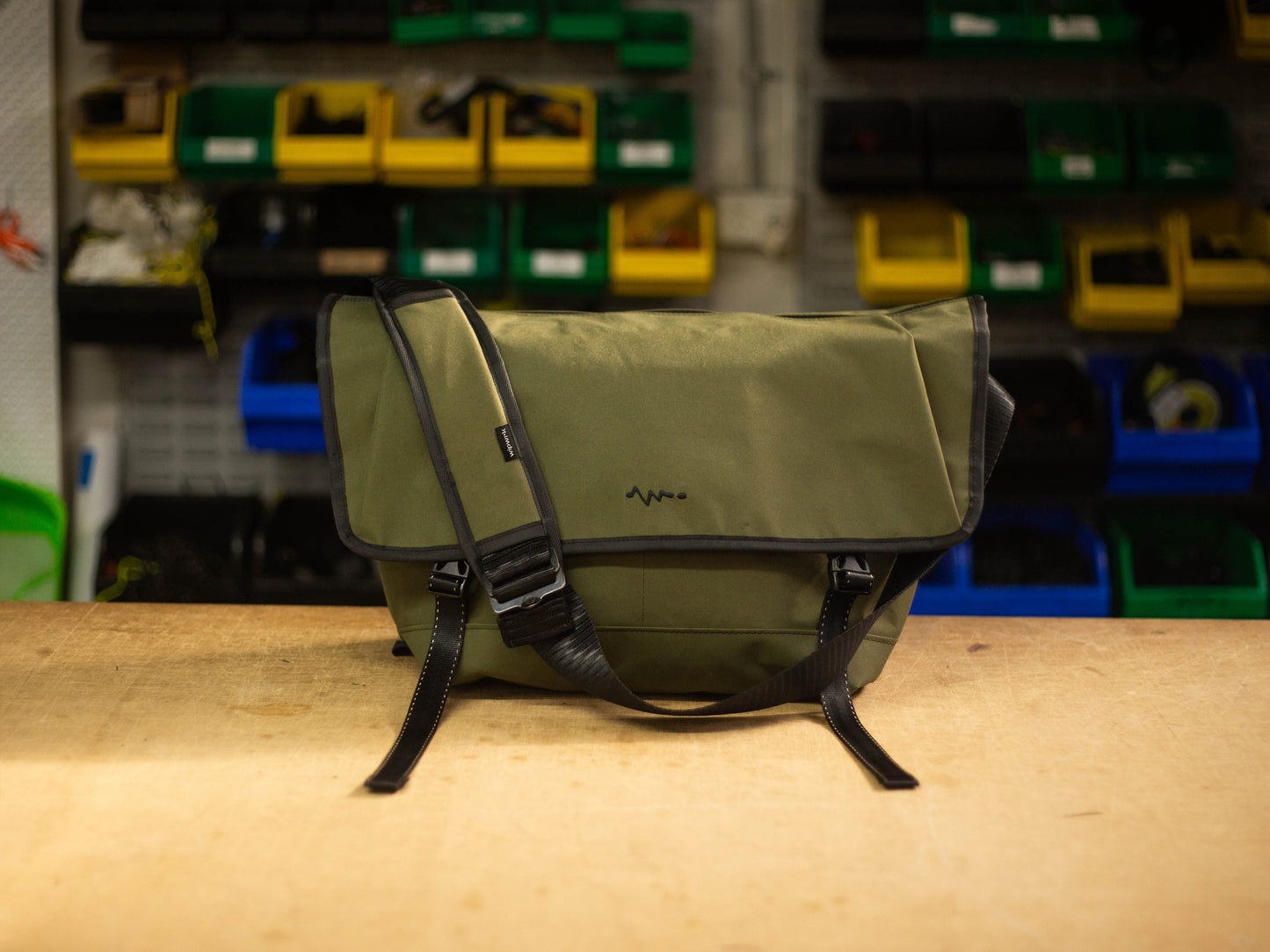 Front view of green wipwrk Mondo messenger bag in brown showing durable exterior, adjustable padded shoulder strap, and quick-release buckles against a workshop backdrop