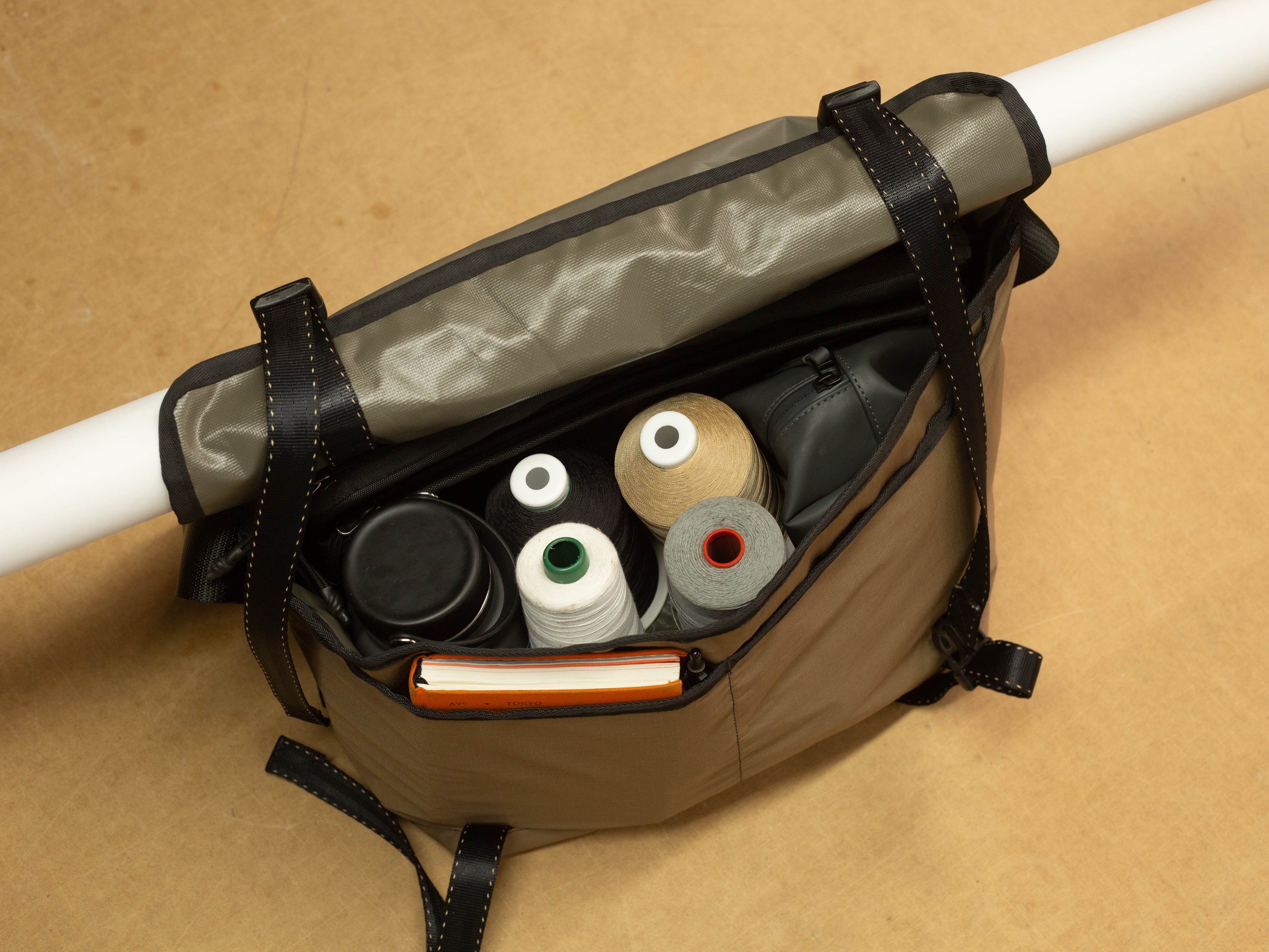 Top-down view of wipwrk Mondo Hauler messenger bag in brown with top flap rolled up and secured by integrated straps holding a rolled document tube, interior packed with sewing thread spools, reusable water bottle, zippered organizer pouch, and notebook